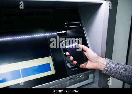 Mann`s Hand mit einem Smartphone mit einem Geldautomaten an der Straße, um etwas Geld abzuheben Stockfoto